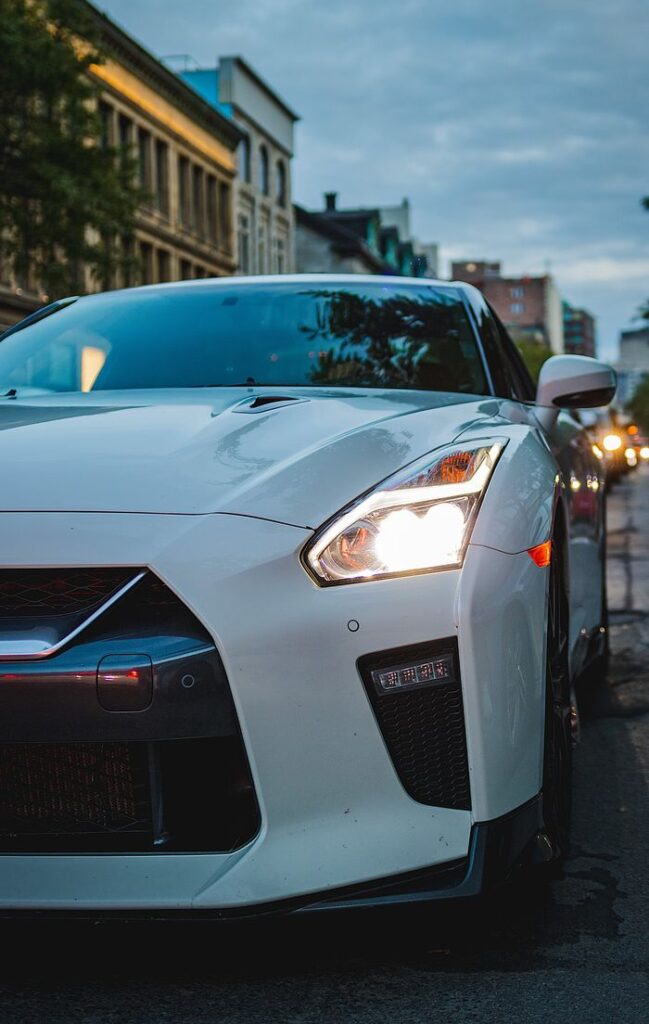 White sports car with headlights on, parked on a city street at dusk.