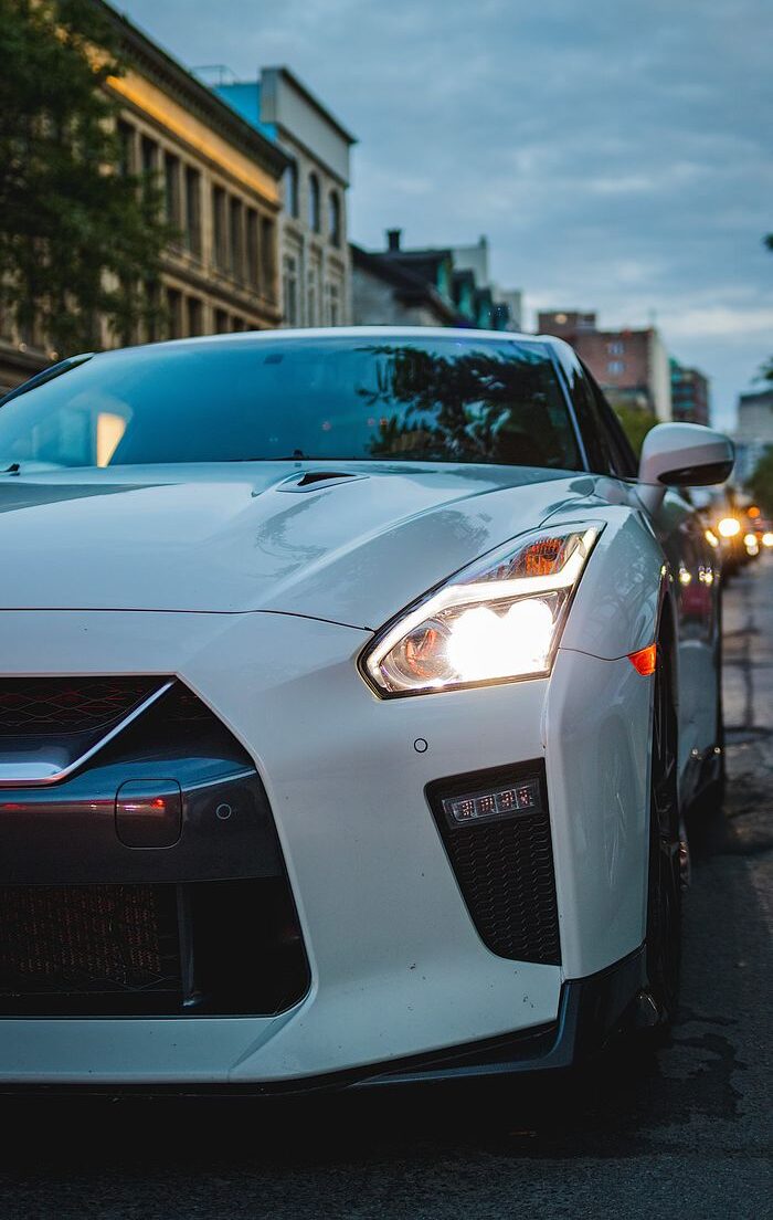 White sports car with headlights on, parked on a city street at dusk.